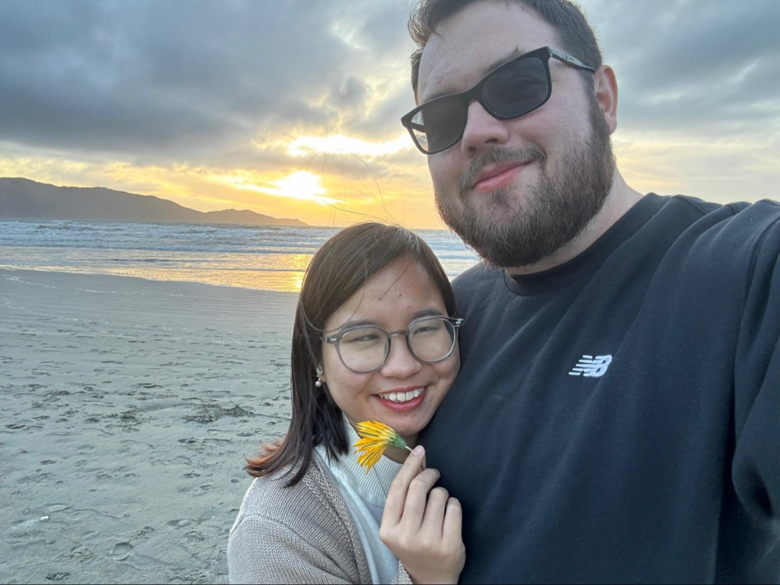 Trang and Robert on a beach at sunset, Trang holding a small yellow flower