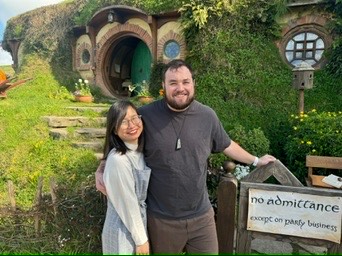 Trang and Robert in front of a round green hobbit hole door at Hobbiton